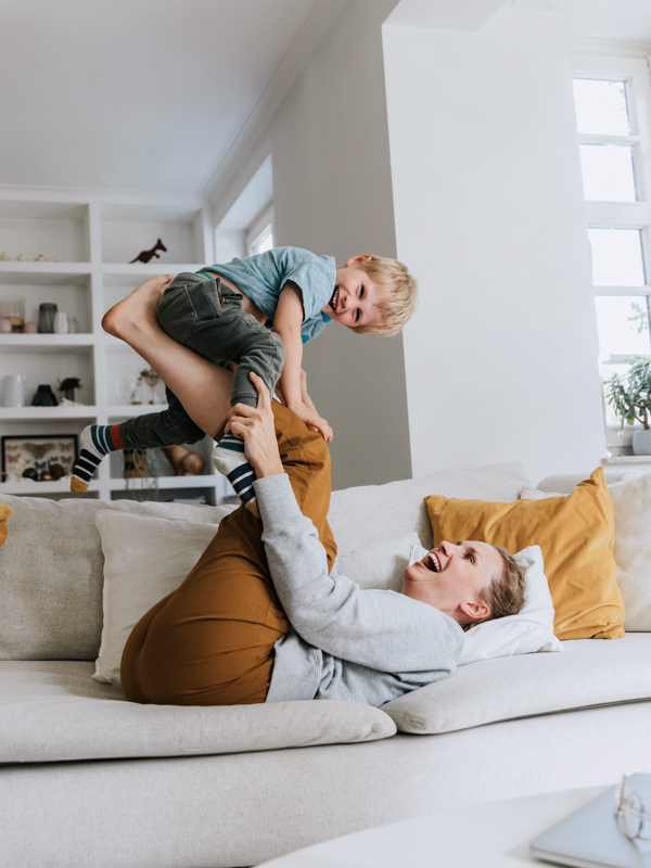Eine Mutter liegt auf der Couch und stemmt spielerisch ihren jungen Sohn mit den Füßen in die Höhe. Beide lachen